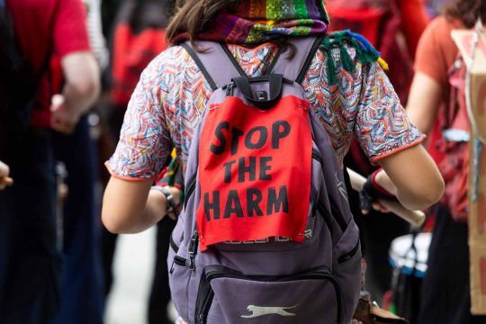 Image shows a person's torso from behind, wearing multi-coloured t-shirt and rucksack with 'Stop the harm' messages on fabric pinned to it.