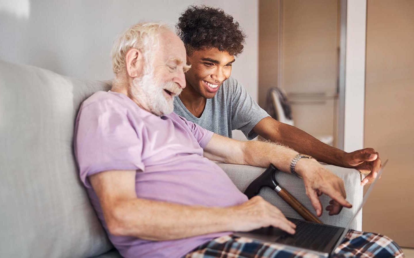 Older person sitting on sofa using laptop and walking stick by their side. Careworker looking at the same laptop, both smiling.