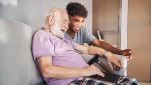 Older person sitting on sofa using laptop and walking stick by their side. Careworker looking at the same laptop, both smiling.