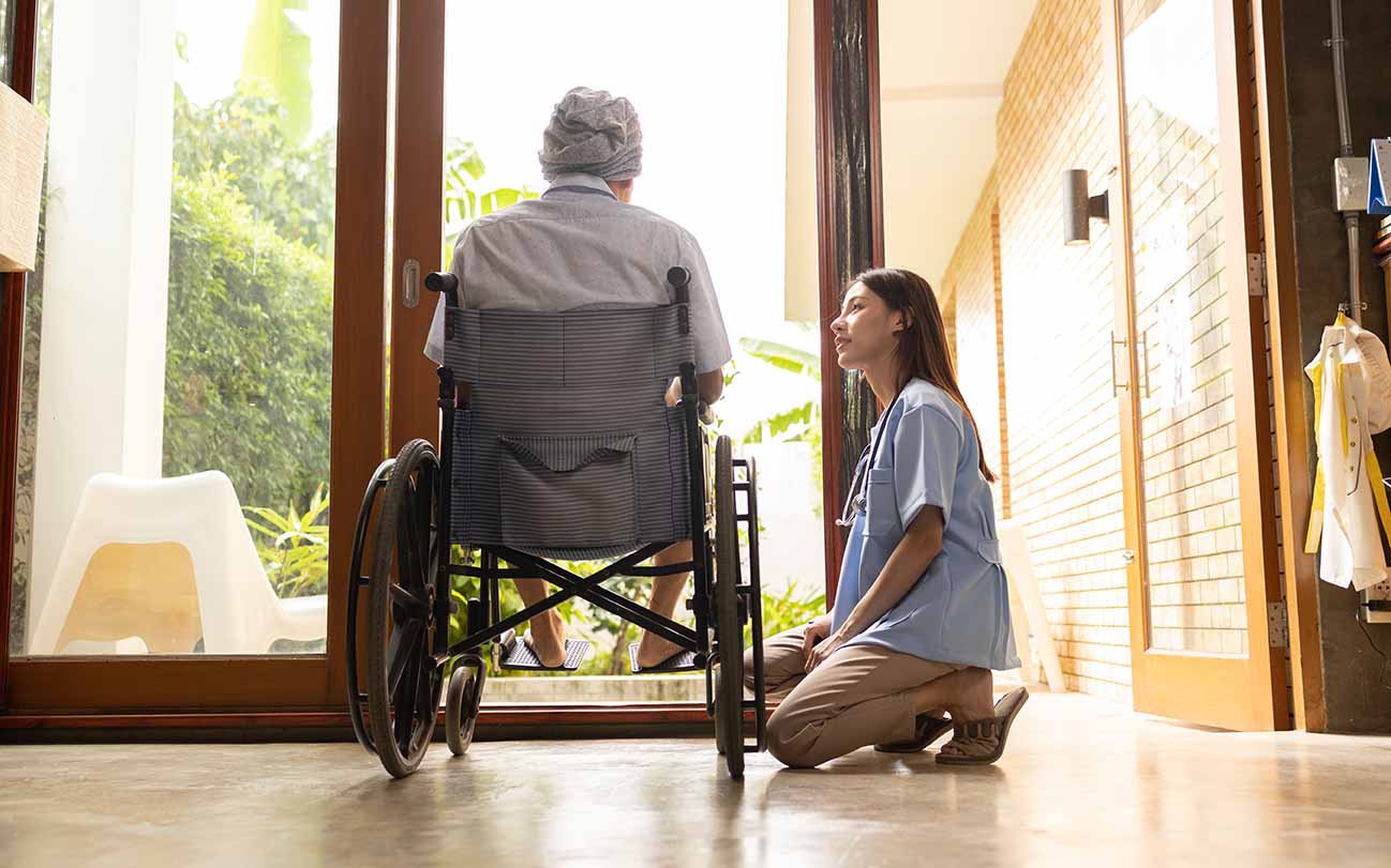 Older person sitting in wheelchair facing away from camera, looking through a large glass door towards a garden, younger person in work clothes kneeling down next to the wheelchair, looking into the older person's face.