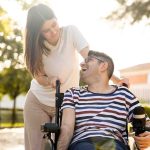 An adult man with a disability sits in an automatic wheelchair with a young woman in a park at sunset.The woman and the man look at each other happily.