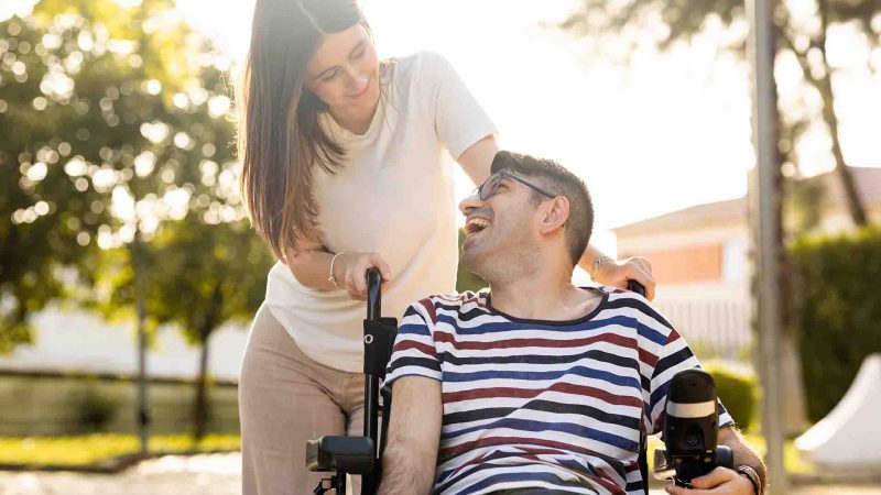 An adult man with a disability sits in an automatic wheelchair with a young woman in a park at sunset.The woman and the man look at each other happily.