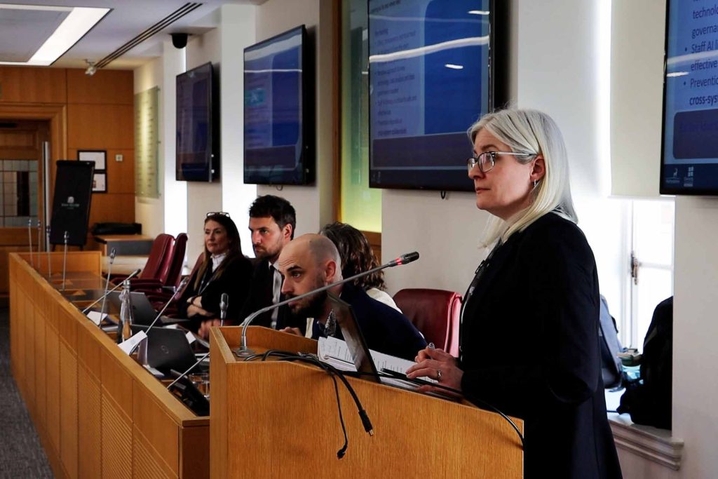 Kate Hamblin standoing at podium with panel of experts sitting behind her.