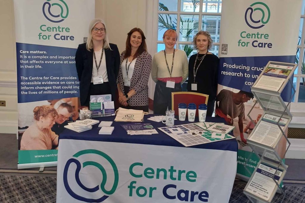 Centre for Care team members posing for a photo at a conference exhibition with Centre for Care- branded merchandise on a table and on poster banners.