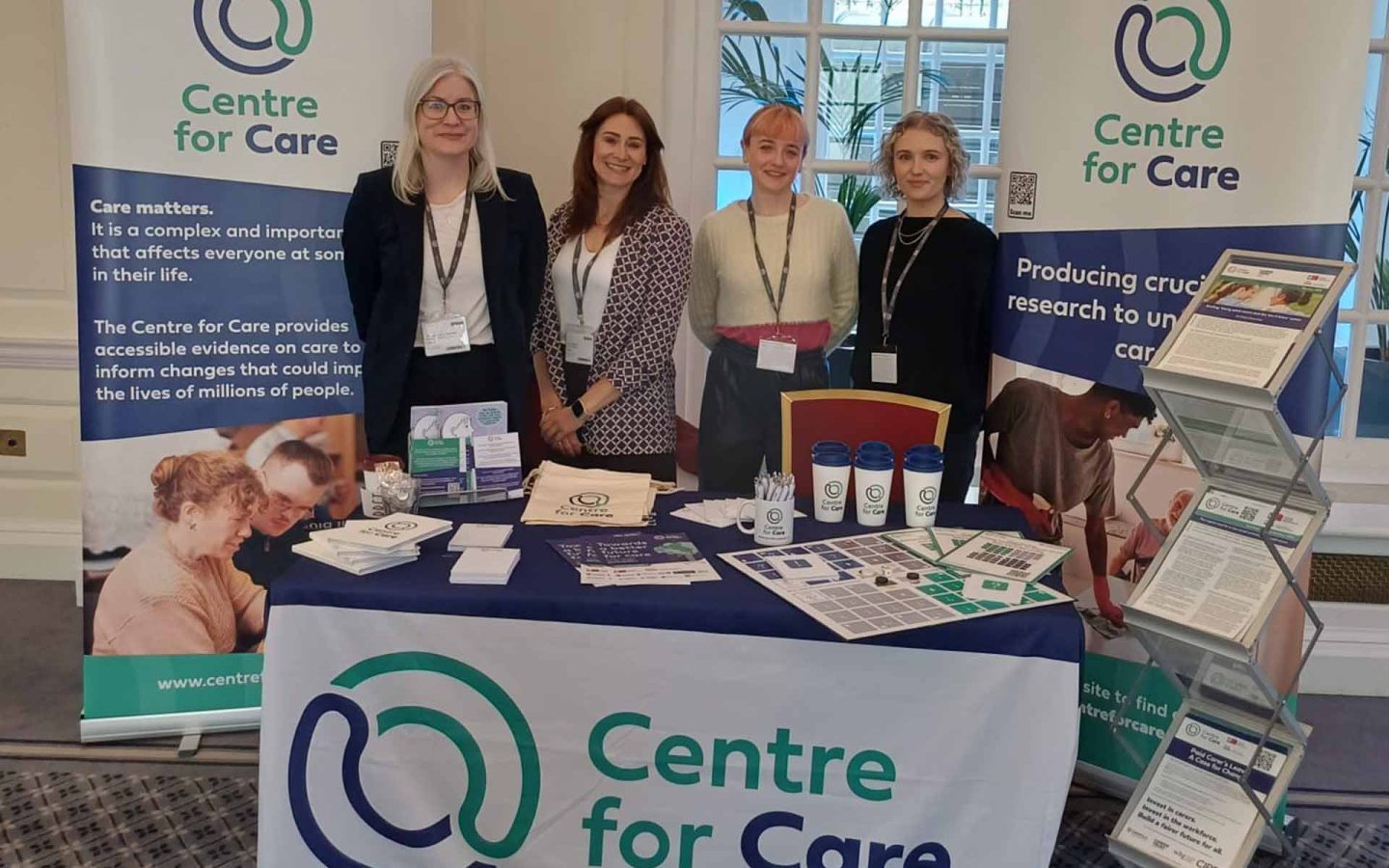 Centre for Care team members posing for a photo at a conference exhibition with Centre for Care- branded merchandise on a table and on poster banners.