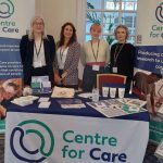 Centre for Care team members posing for a photo at a conference exhibition with Centre for Care- branded merchandise on a table and on poster banners.