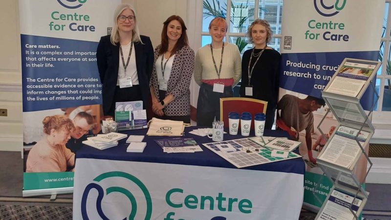 Centre for Care team members posing for a photo at a conference exhibition with Centre for Care- branded merchandise on a table and on poster banners.