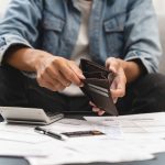Person lookinng through empty wallet with calculator and paperwork on a table in front of them