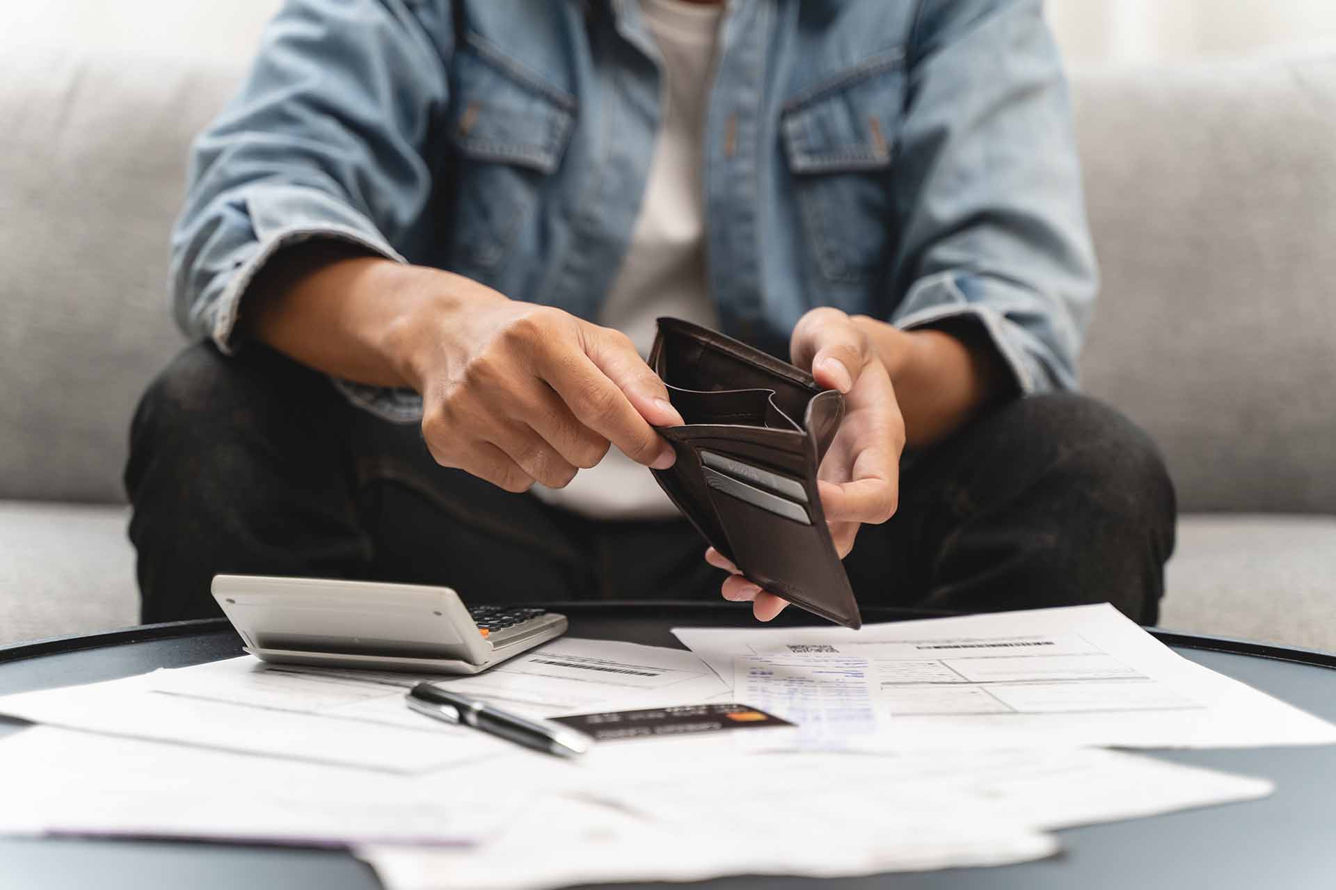 Person lookinng through empty wallet with calculator and paperwork on a table in front of them