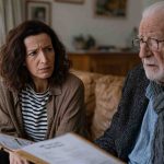 adult daughter sitting across from her elderly father at his home, a folder of care facility options between them, both with tense thoughtful expressions, aging parent