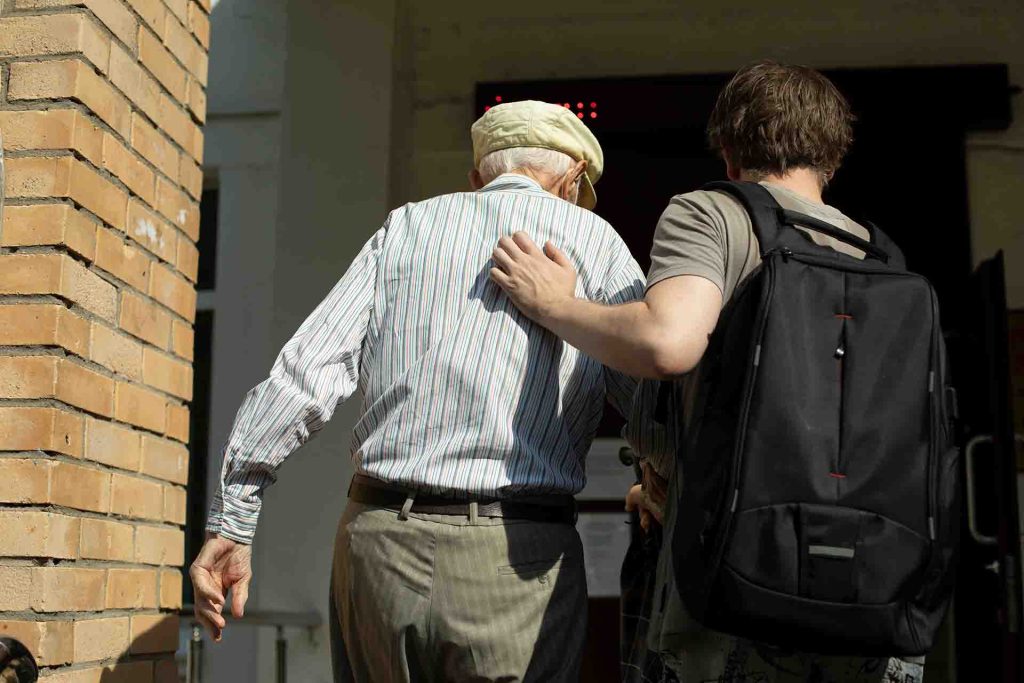 Younger man helping older man to walk through an entrace way, younger man is wearing a backpack and they are both walking away from the camera