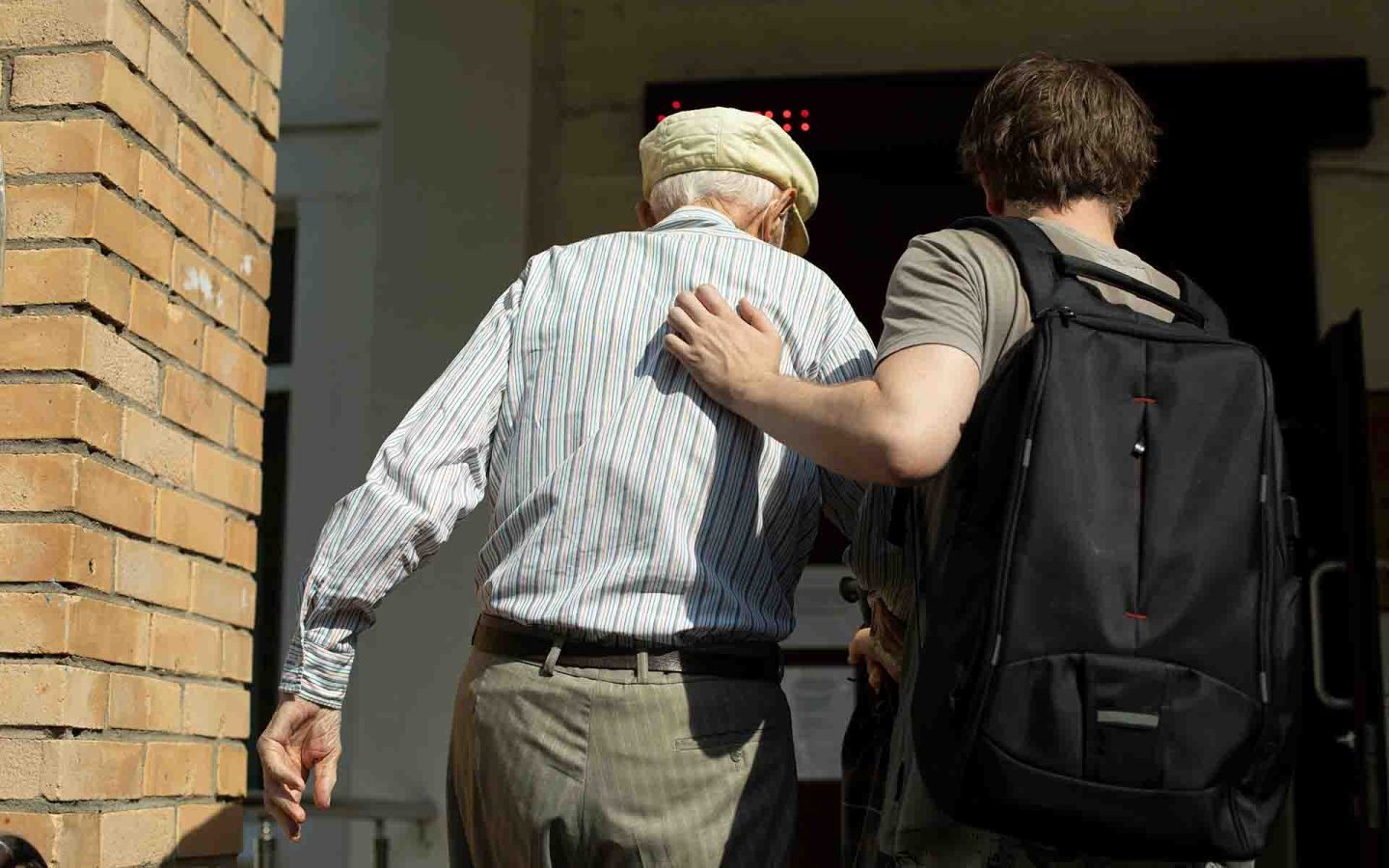 Younger man helping older man to walk through an entrace way, younger man is wearing a backpack and they are both walking away from the camera