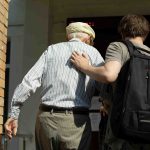Younger man helping older man to walk through an entrace way, younger man is wearing a backpack and they are both walking away from the camera