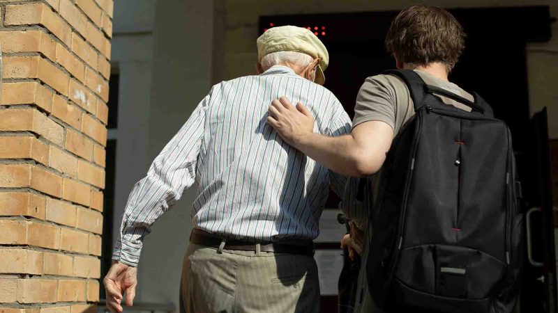 Younger man helping older man to walk through an entrace way, younger man is wearing a backpack and they are both walking away from the camera