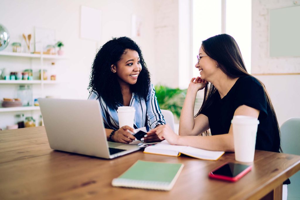 two people sit at desk with a laptop and paperwork, they are talking and smiling