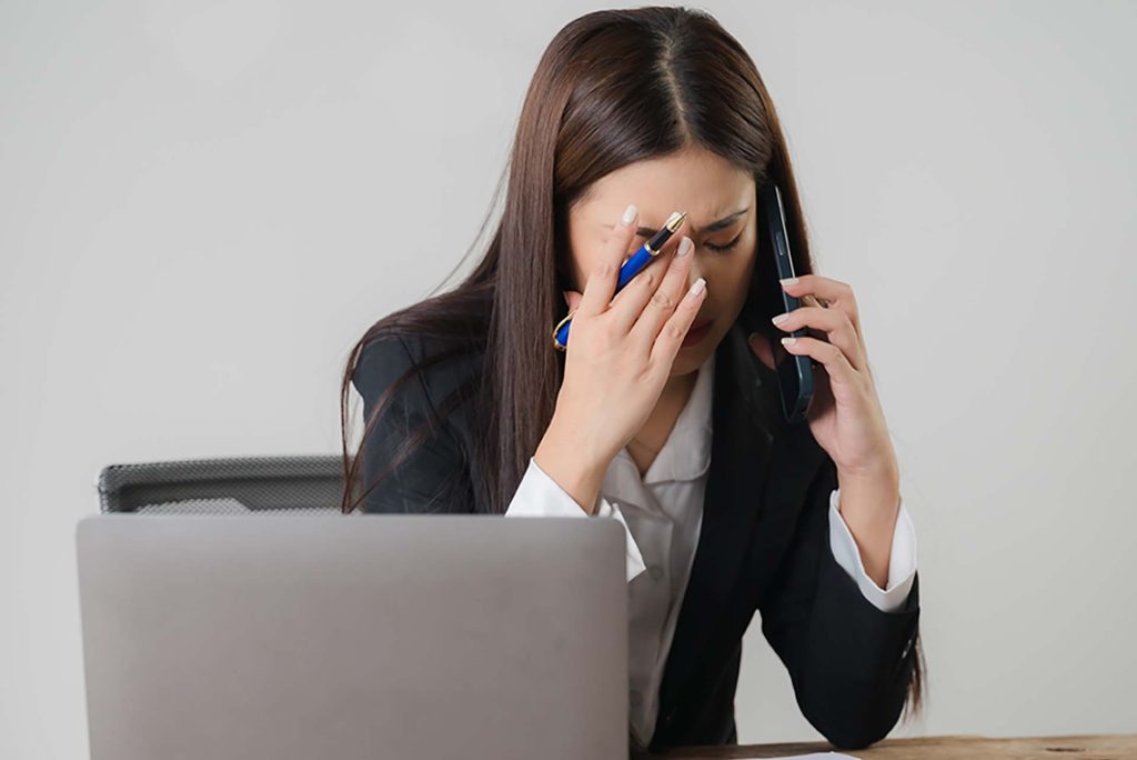 woman sitting at desk in front of laptop using mobile phone and looking worried