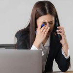 woman sitting at desk in front of laptop using mobile phone and looking worried