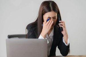 woman sitting at desk in front of laptop using mobile phone and looking worried