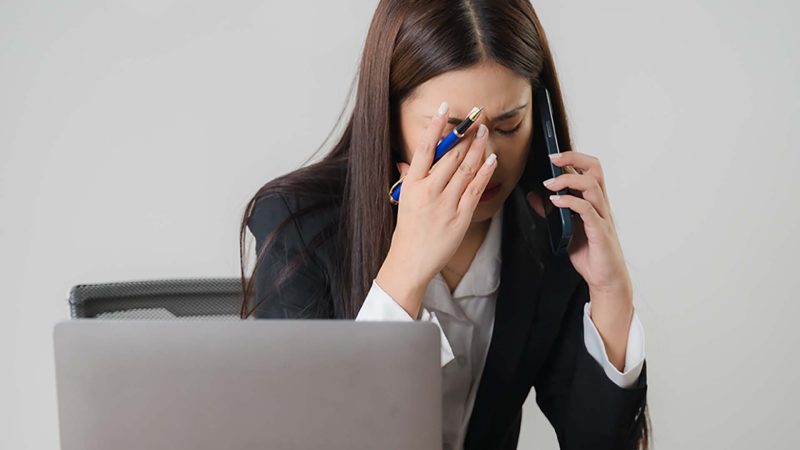 woman sitting at desk in front of laptop using mobile phone and looking worried